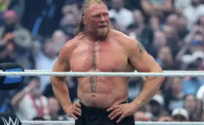 Muscular male wrestler stands with hands on hips in a ring, sweaty with a cheering crowd behind him. Wrestler in black shorts, long hair and chest tattoo, posing inside the ring. Athletic man with a vertical chest tattoo, standing shirtless inside a wrestling ring as spectators look on. Confident wrestler wearing black trunks, arms akimbo, ropes across midsection with blurred audience. Strong-built performer in a wrestling arena, focusing forward with crowd in the background.