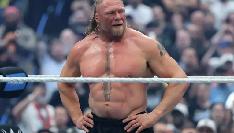 Muscular male wrestler stands with hands on hips in a ring, sweaty with a cheering crowd behind him. Wrestler in black shorts, long hair and chest tattoo, posing inside the ring. Athletic man with a vertical chest tattoo, standing shirtless inside a wrestling ring as spectators look on. Confident wrestler wearing black trunks, arms akimbo, ropes across midsection with blurred audience. Strong-built performer in a wrestling arena, focusing forward with crowd in the background.