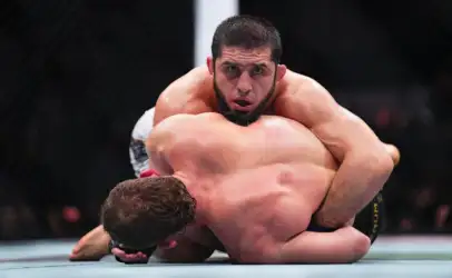 Two male fighters grapple on the mat inside a cage, with one on top in a controlling position during a match.