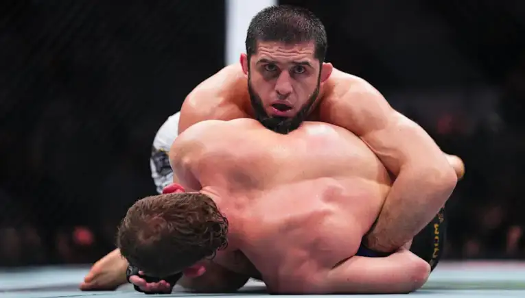 Two male fighters grapple on the mat inside a cage, with one on top in a controlling position during a match.