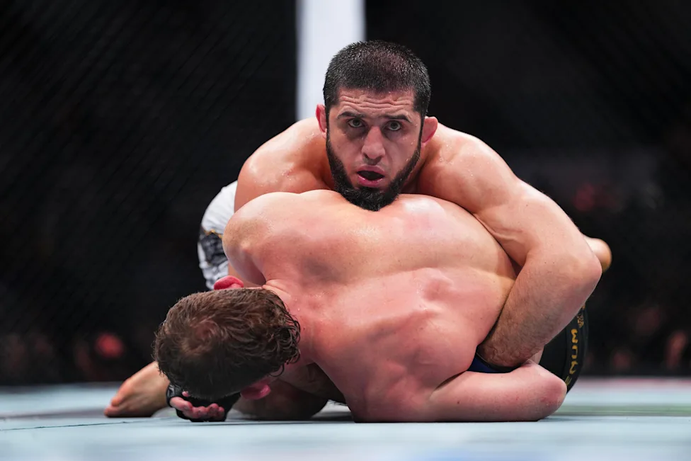 ff651862839e1dd30df6cdb7ce32c101 - AfroCombat Two male fighters grapple on the mat inside a cage, with one on top in a controlling position during a match.