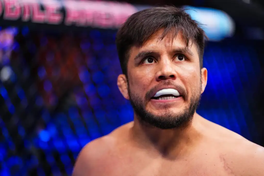 Close-up of a shirtless man looking upward, teeth visible, inside a blue-lit arena surrounded by a cage fence.
