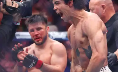 Shirtless boxer shouting in celebration in a boxing ring, as a cameraman films from the left.