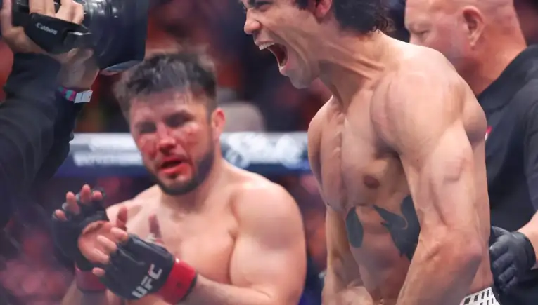 Shirtless boxer shouting in celebration in a boxing ring, as a cameraman films from the left.