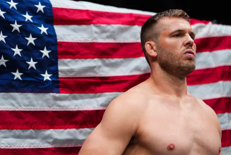 Shirtless male athlete with short hair standing in front of a waving American flag.