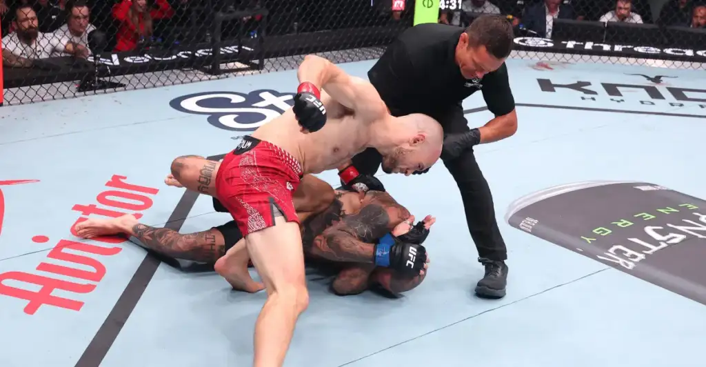 Two mixed martial artists grapple on the mat while a referee watches nearby inside an arena Octagon/cage.
