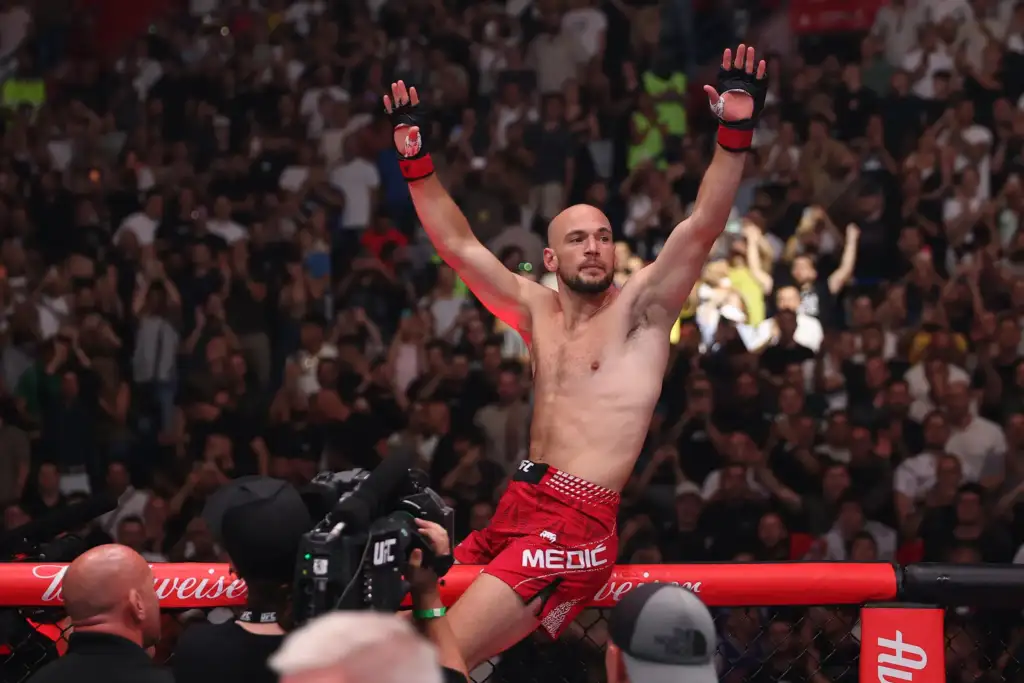 MMA fighter with red shorts raises both arms in victory inside the cage as a cheering crowd watches behind him.