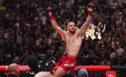 MMA fighter with red shorts raises both arms in victory inside the cage as a cheering crowd watches behind him.