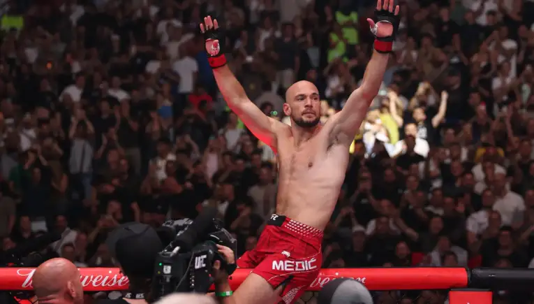 MMA fighter with red shorts raises both arms in victory inside the cage as a cheering crowd watches behind him.