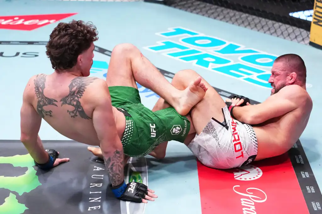 Two shirtless mixed martial artists grappling on a UFC-style mat, one with a large back tattoo and green shorts, the other in white shorts.
