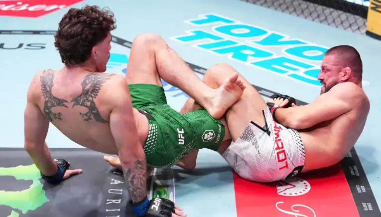 Two shirtless mixed martial artists grappling on a UFC-style mat, one with a large back tattoo and green shorts, the other in white shorts.