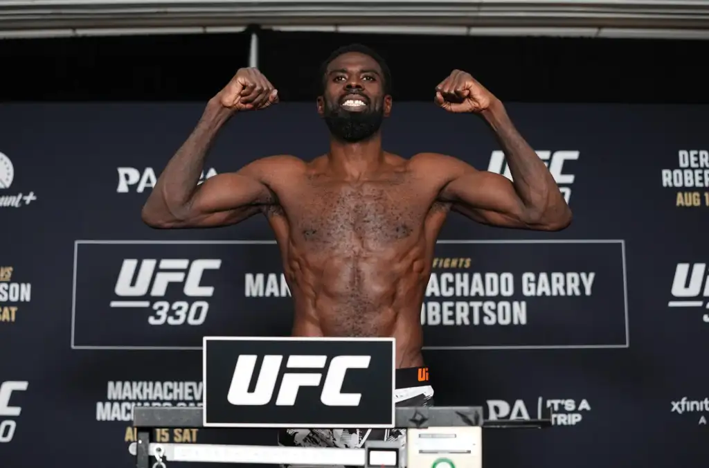 Male UFC fighter on a weigh-in stage, shirtless, flexing arms with a UFC backdrop behind him.