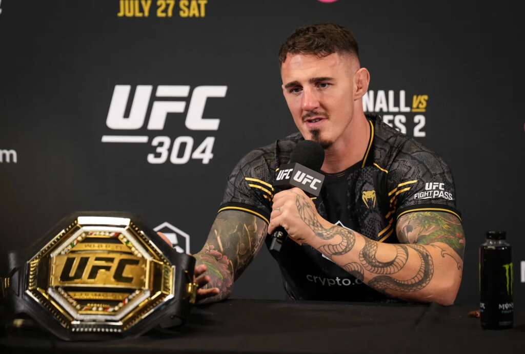 UFC fighter speaks into a microphone at a press conference with a championship belt on the table nearby.