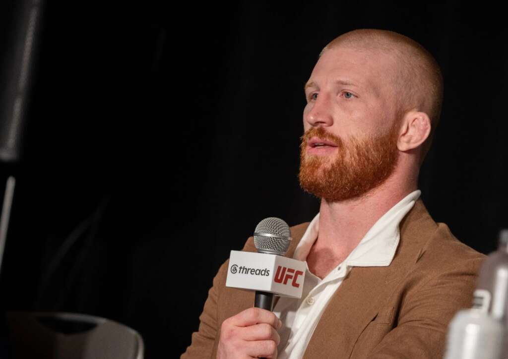 Bearded man in a brown blazer speaks into a handheld microphone with UFC and Threads logos visible on the mic tag