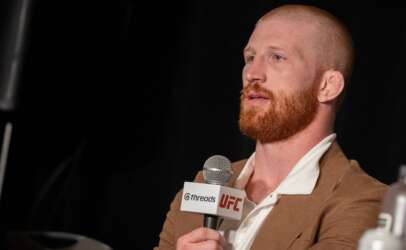 Bearded man in a brown blazer speaks into a handheld microphone with UFC and Threads logos visible on the mic tag
