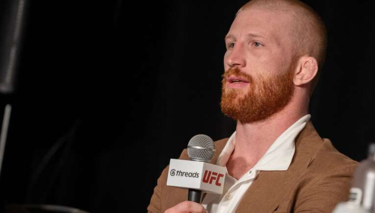 Bearded man in a brown blazer speaks into a handheld microphone with UFC and Threads logos visible on the mic tag