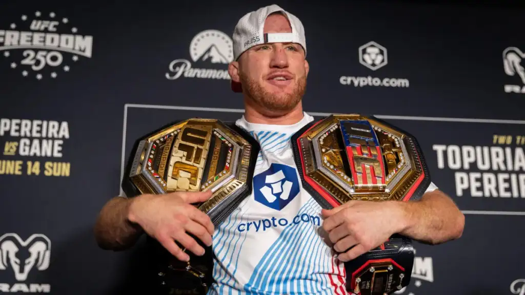 UFC fighter posing with two championship belts over shoulders at a press backdrop, crypto.com logo on shirt visible.