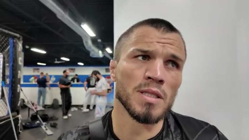 umar-nurmagomedov-merab-dvalishvili-raf-georgia-reaction-1600x900-1 - AfroCombat Close-up of a man with short hair and stubble looking to the side in a gym, with people training in the background.