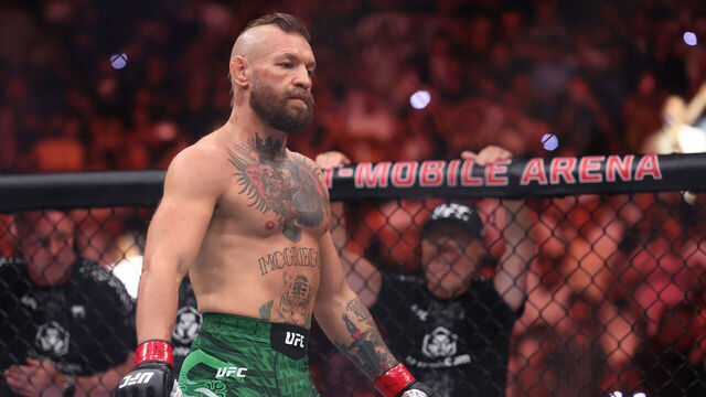 UFC fighter with a beard and tattoos standing inside the octagon wearing green shorts and red gloves, arena crowd in the background.