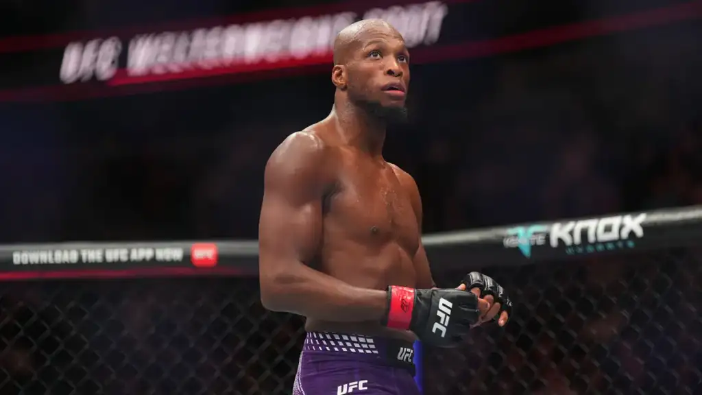 Shirtless male mixed martial artist stands inside the UFC octagon wearing purple shorts and black gloves, looking upward with a focused expression.