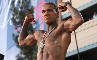 Muscular, tattooed man flexing his biceps on stage at an outdoor event, wearing a necklace and watch.