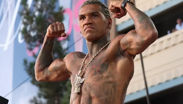 Muscular, tattooed man flexing his biceps on stage at an outdoor event, wearing a necklace and watch.