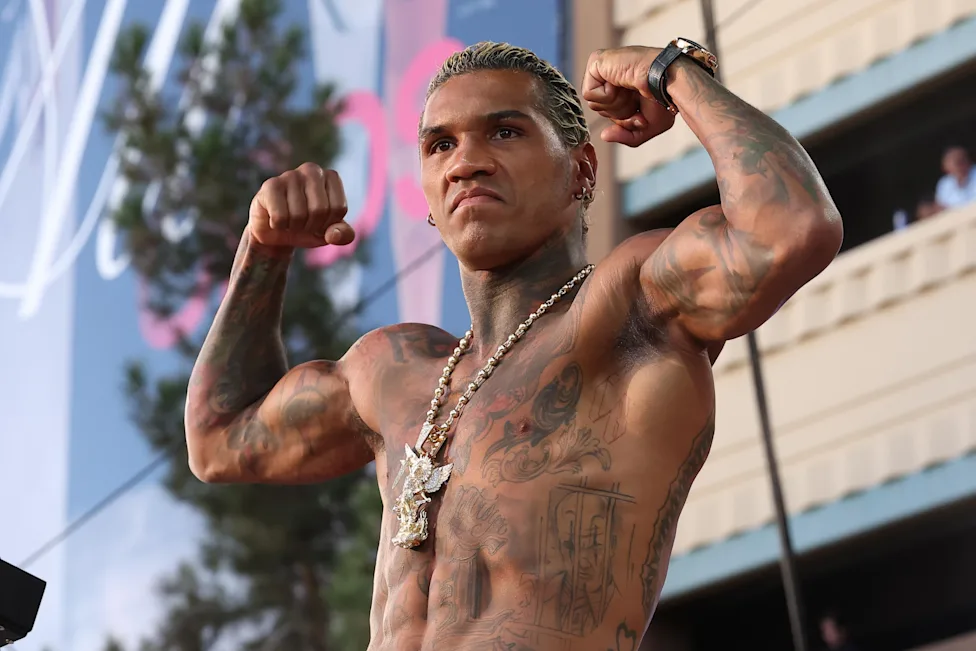 03346dd01082db90e1ed666c41fbbd85 - AfroCombat Muscular, tattooed man flexing his biceps on stage at an outdoor event, wearing a necklace and watch.