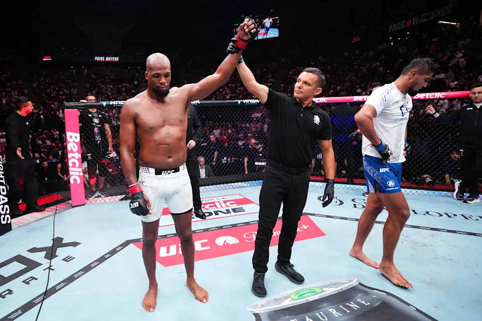 0419df4839b6903e14006fb99287324e - AfroCombat UFC fighter in white shorts with fist raised by referee, standing in the octagon as the opponent in blue looks down in defeat, crowd in the background.