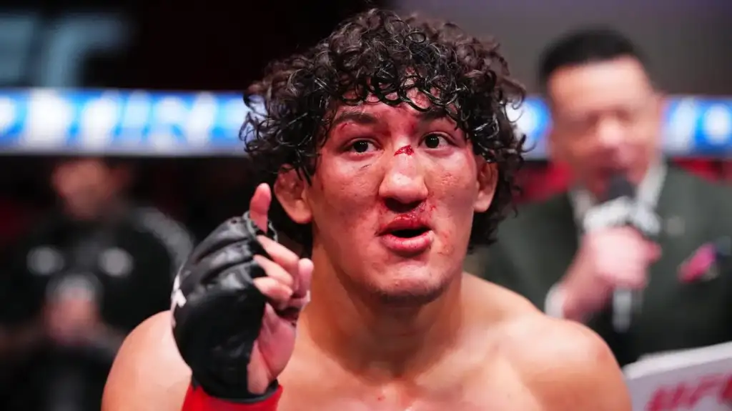 Bloodied mixed martial arts fighter with curly hair speaks to camera in a ring, reporter visible in the background.