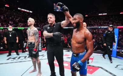 UFC victory moment: a muscular fighter in blue shorts and gloves stands with a raised arm as the referee signals the win, while his tattooed opponent in black shorts watches inside the octagon.
