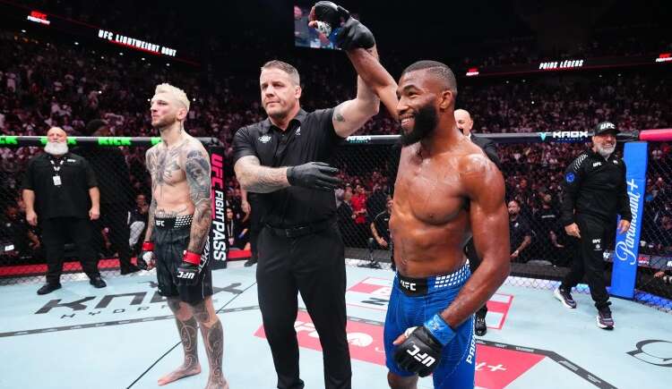 UFC victory moment: a muscular fighter in blue shorts and gloves stands with a raised arm as the referee signals the win, while his tattooed opponent in black shorts watches inside the octagon.