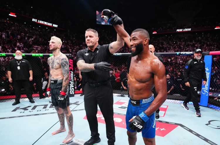 UFC victory moment: a muscular fighter in blue shorts and gloves stands with a raised arm as the referee signals the win, while his tattooed opponent in black shorts watches inside the octagon.