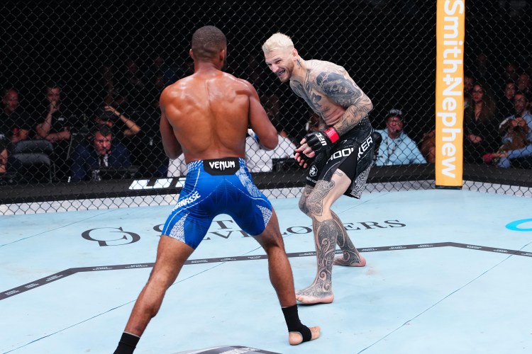 Two male mixed martial artists face off inside a cage, one with blue shorts delivering a stance while the tattoed fighter in black shorts advances aggressively. Audience watches from the stands.
