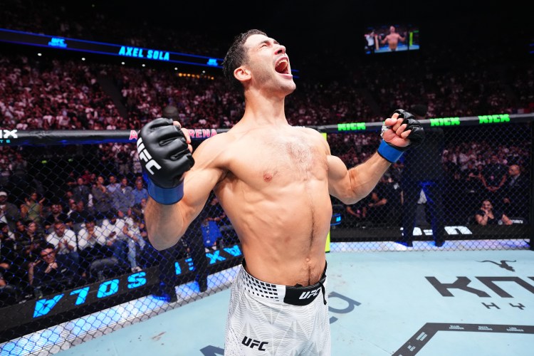 UFC fighter in the octagon celebrates with fists raised and mouth open in victory as the crowd cheers in the background