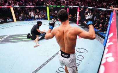 UFC fighter seen from behind raises his gloved hands inside the octagon, opponent on the mat and referee nearby.