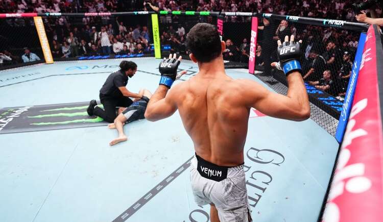 UFC fighter seen from behind raises his gloved hands inside the octagon, opponent on the mat and referee nearby.