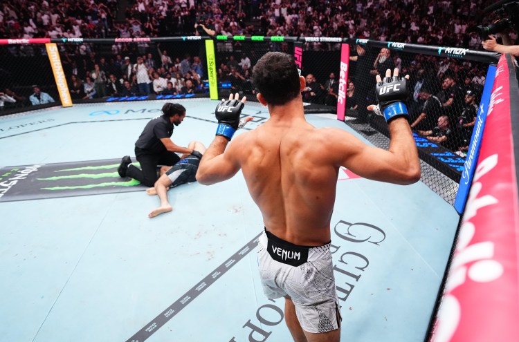 UFC fighter seen from behind raises his gloved hands inside the octagon, opponent on the mat and referee nearby.