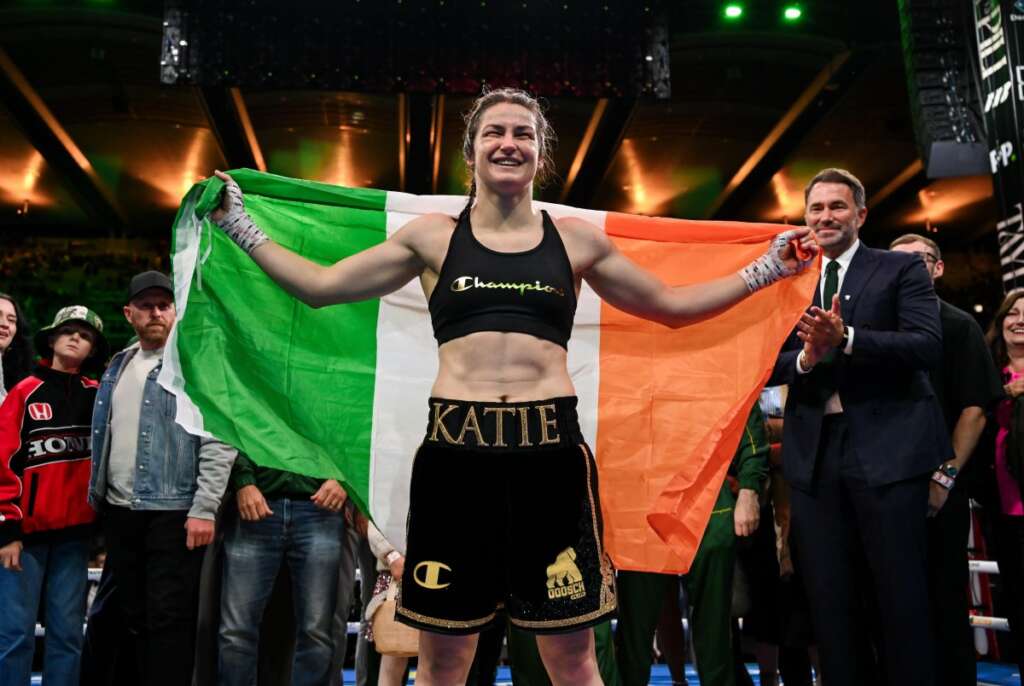 Female boxer celebrating in the ring, arms outstretched with an Italian flag behind her, wearing black Champion top and 'KATIE' shorts belt.