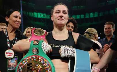 Female boxer in victory pose holding a colorful World Boxing belt amid cheering crowd; WBO badge visible on lanyard and gloves strapped.