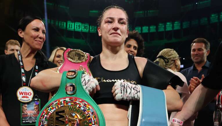 Female boxer in victory pose holding a colorful World Boxing belt amid cheering crowd; WBO badge visible on lanyard and gloves strapped.