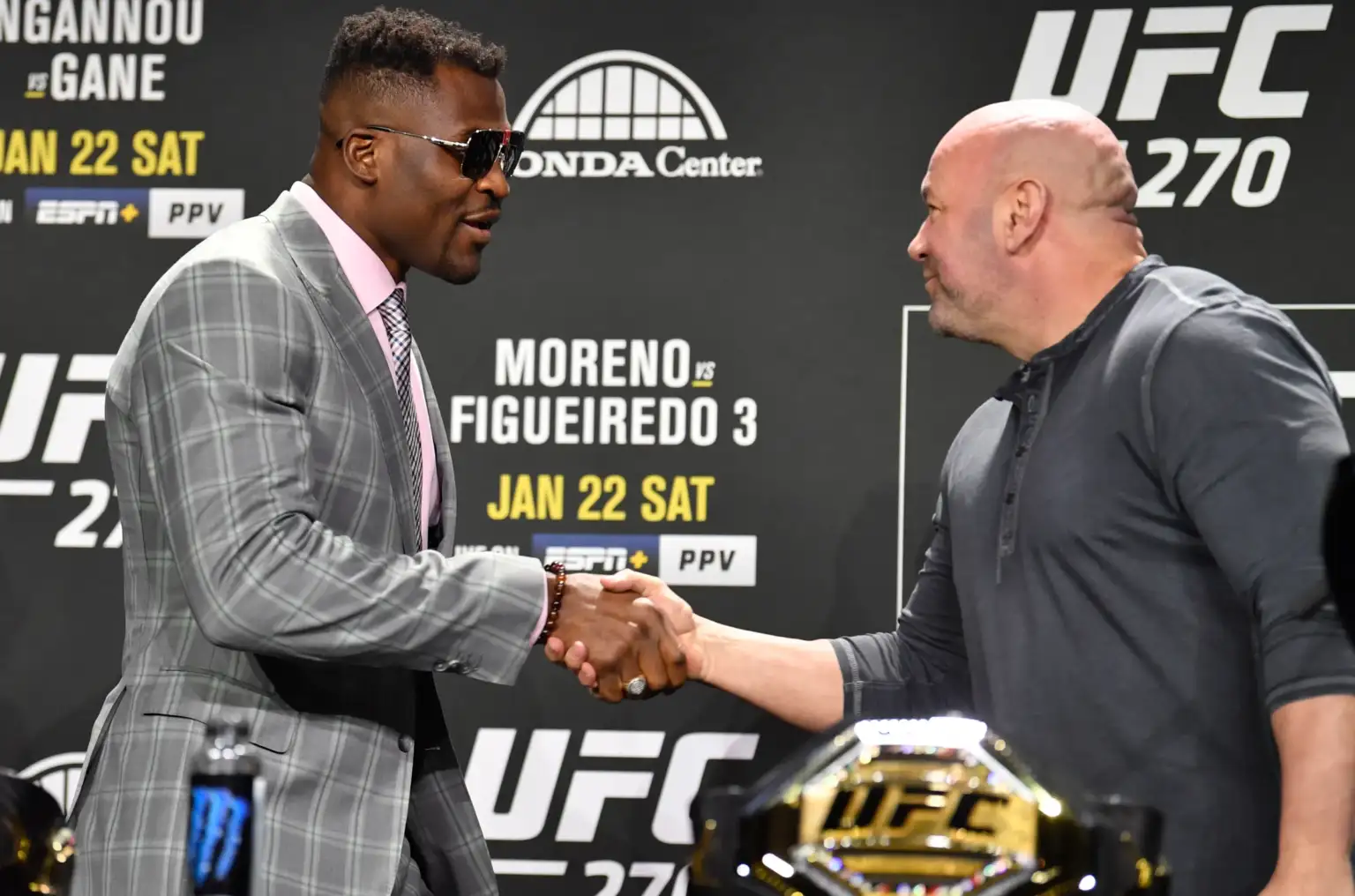 Two men shake hands at a UFC weigh‑in, one in a gray checkered suit and pink shirt, the other in a dark polo, with a UFC belt in the foreground.