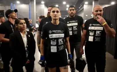 A group of athletes in black shirts with sponsor logos, standing backstage; central female fighter wearing blue gloves looks ahead with others around her.