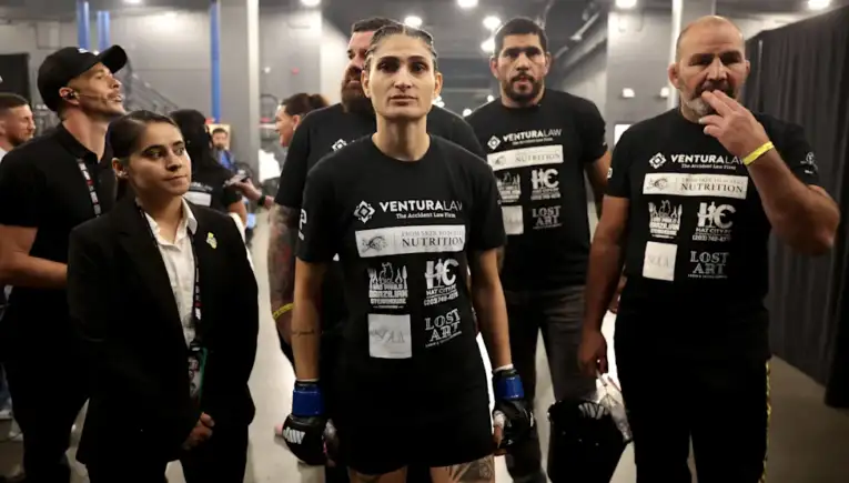 A group of athletes in black shirts with sponsor logos, standing backstage; central female fighter wearing blue gloves looks ahead with others around her.