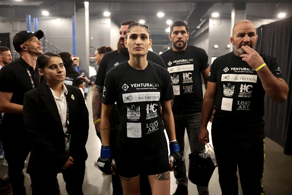 A group of athletes in black shirts with sponsor logos, standing backstage; central female fighter wearing blue gloves looks ahead with others around her.