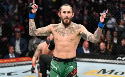 Tattooed MMA fighter with green shorts raises both arms in victory inside the UFC cage.