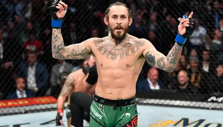 Tattooed MMA fighter with green shorts raises both arms in victory inside the UFC cage.