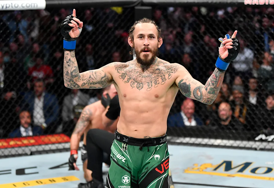 Tattooed MMA fighter with blue gloves raises both arms in victory inside the octagon.