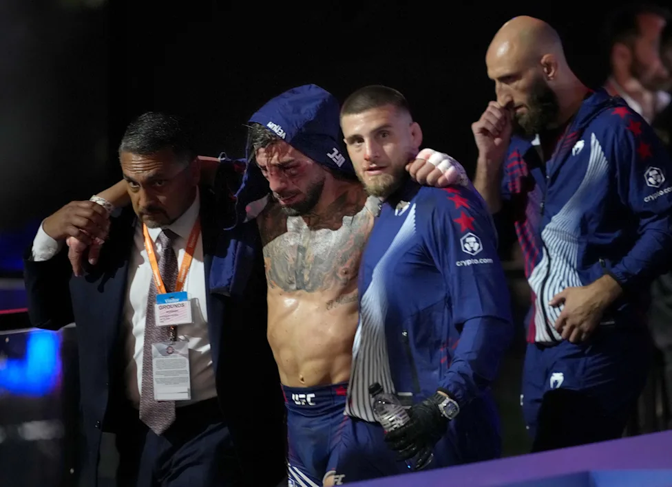 Bruised fighter wearing blue headgear is helped offstage by teammates in blue uniforms and a suited official backstage after a match in a dim arena.