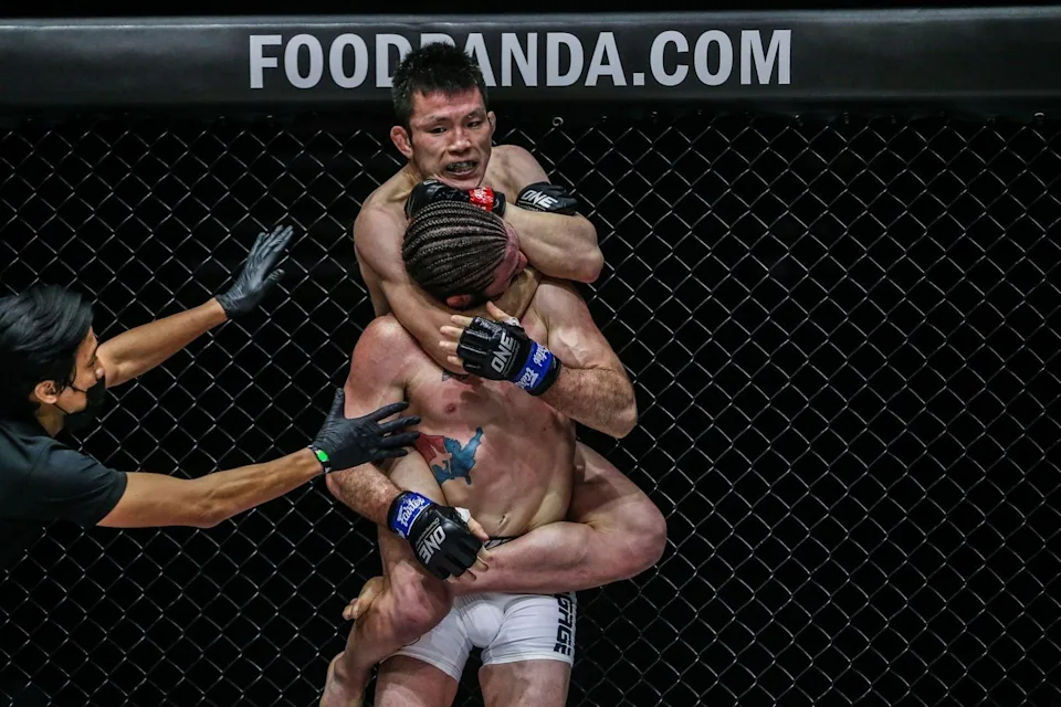 4e74ae638f446a15090b1c306078db79 - AfroCombat Two MMA fighters grapple in a tight clinch inside a chain-link cage while a referee watches from the left; banner above reads FOODANDA.COM.