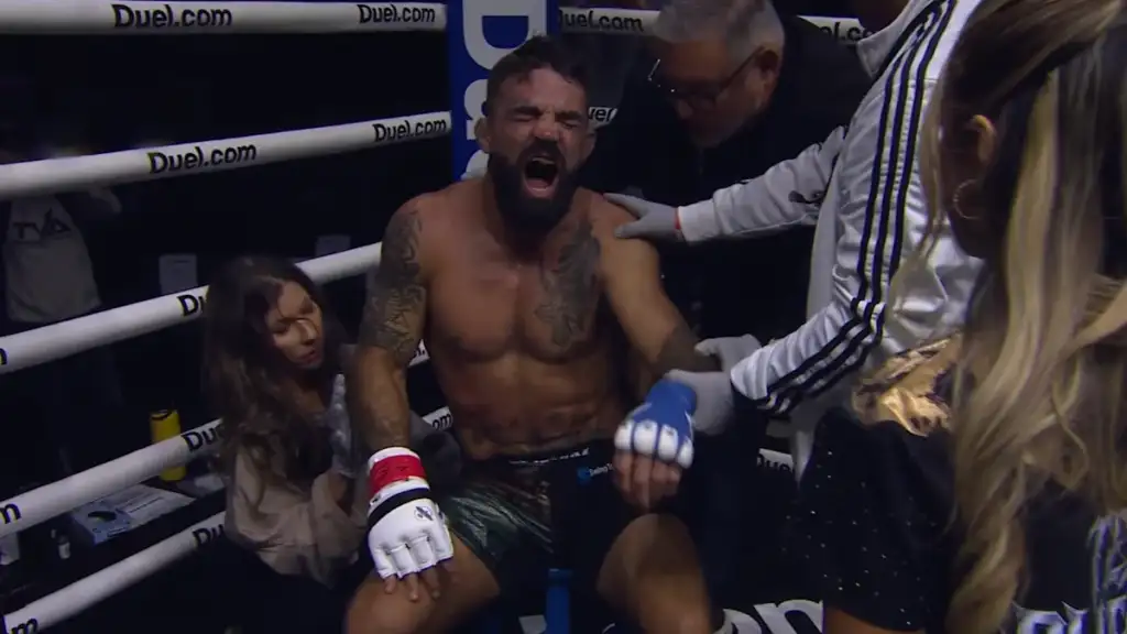 Boxer sits in the corner of a ring, eyes closed and mouth open in a loud yell, surrounded by staff and fans.